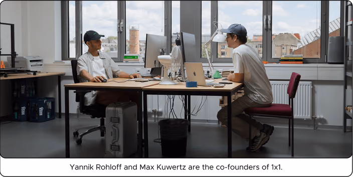 Yannik and Max work together at their desk in a bright office with city views visible through large windows.