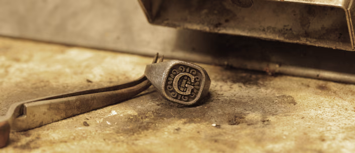 A signet ring engraved with an ornate letter G lies next to pliers on a worn goldsmith's bench.